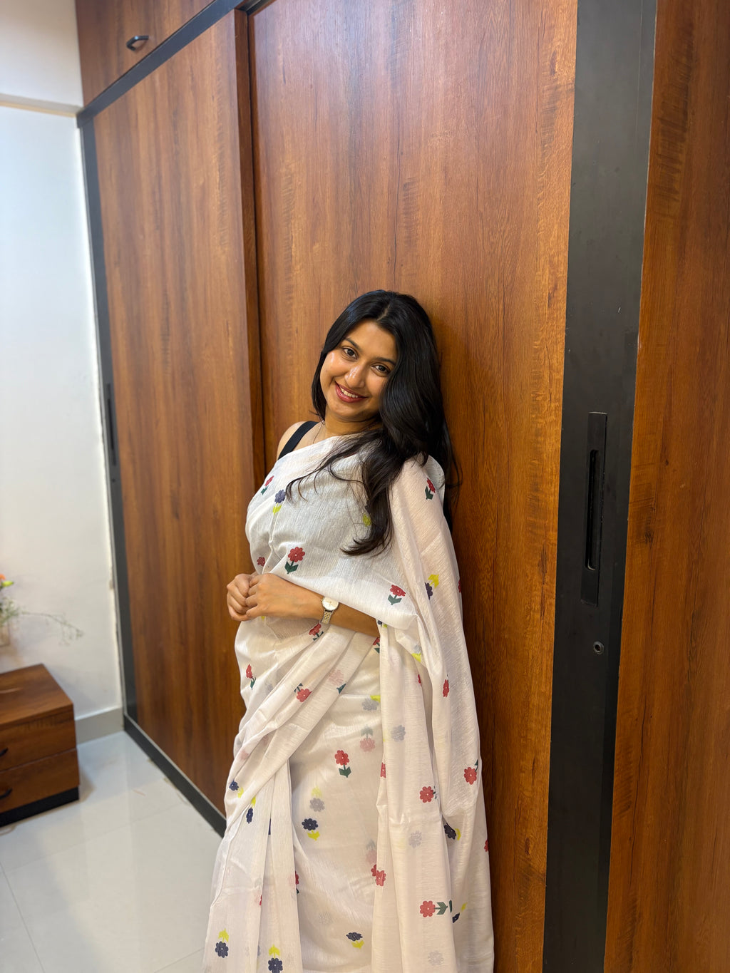 Woman in a white saree with floral patterns standing in front of a wooden wardrobe. Buy cotton white saree.