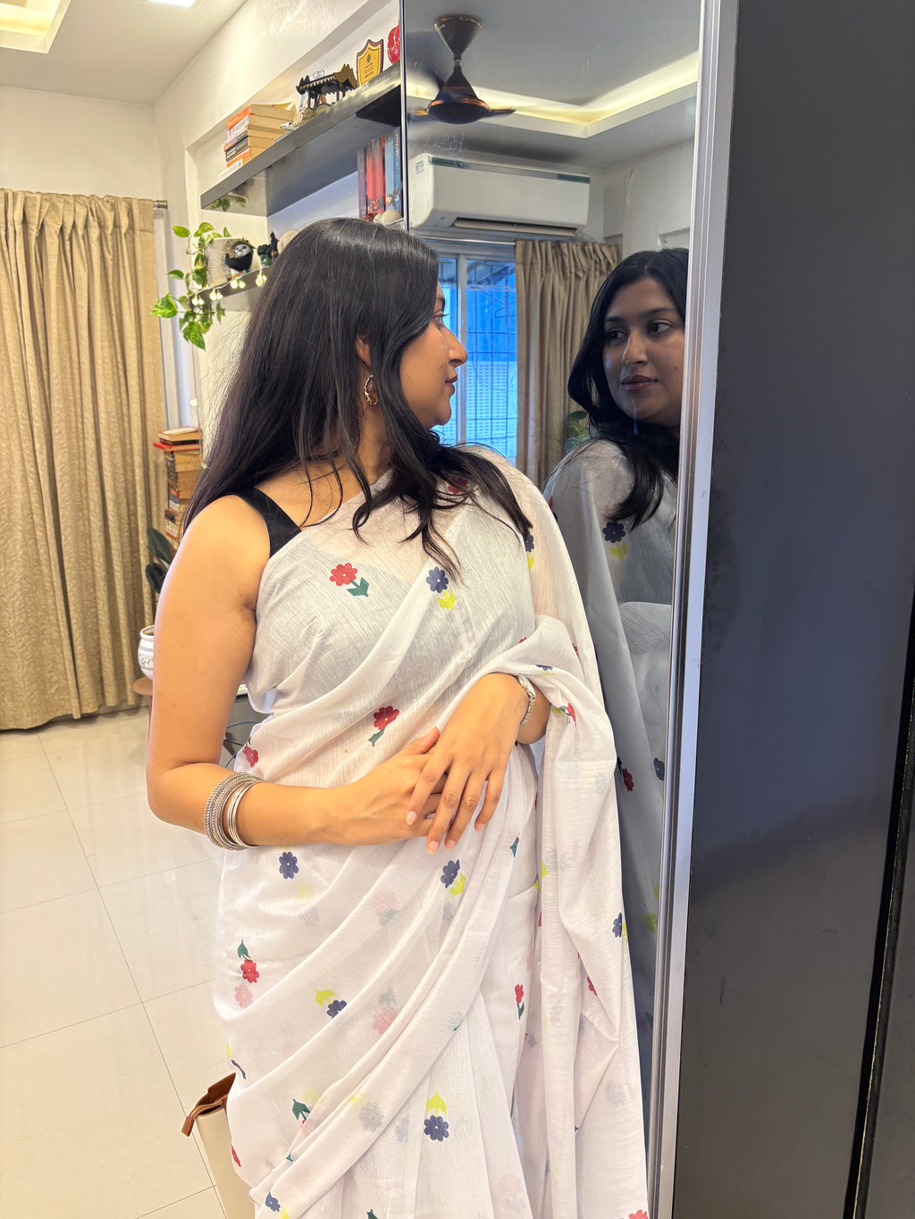 Woman in a white saree with floral patterns standing in front of a mirror. Buy cotton saree.