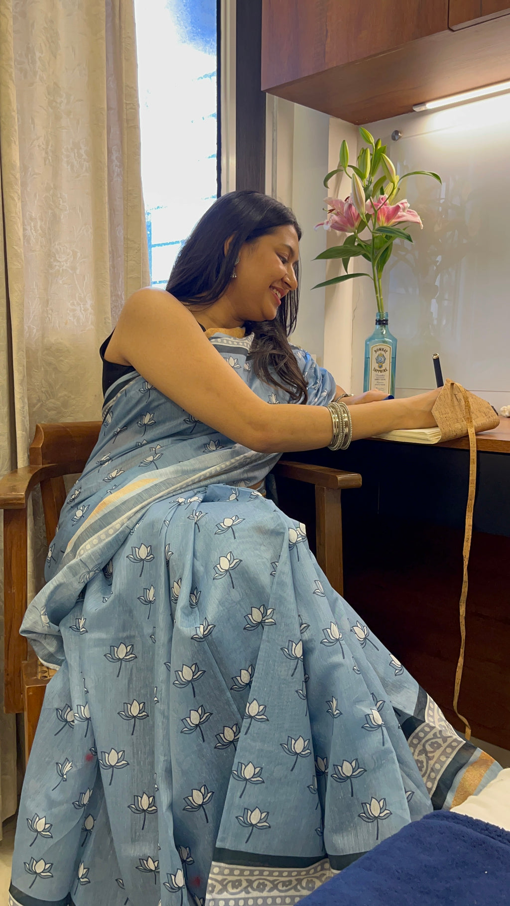 Woman in a blue saree sitting at a desk with a vase of flowers in the background. Women in cotton sari, blue colour, lotus print saree. Lotus print sari. 